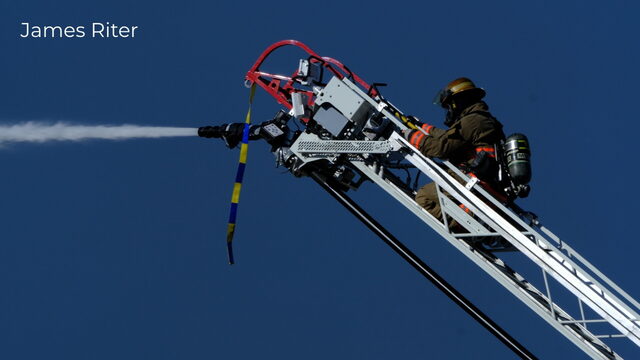 A firefighter directing a stream of water from an aerial ladder against a blue sky, photographed by James Riter
