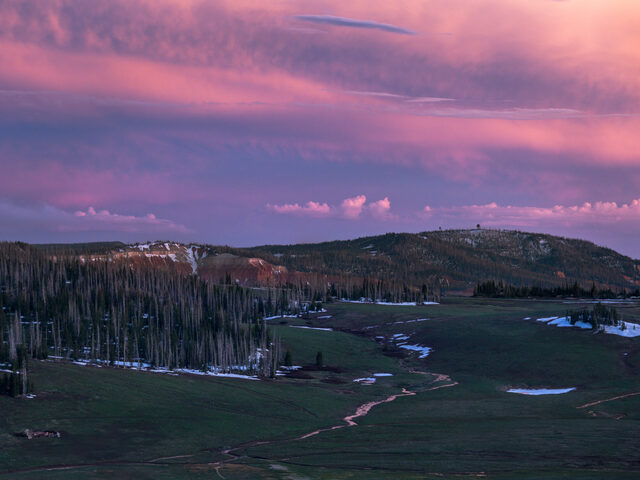 Pink and purple sunset clouds above a mountain meadow, scattered snow, and a winding trail